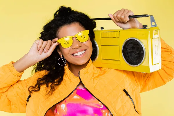Fröhliche afrikanisch-amerikanische junge Frau mit Sonnenbrille und Verkaufsschriftzug hält Boombox isoliert auf gelb — Stockfoto