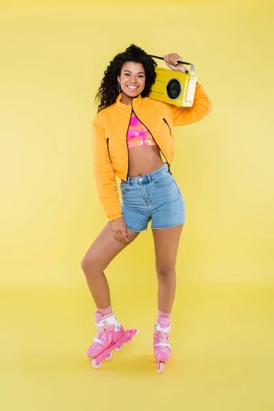 Full length of happy african american woman on roller skates holding retro boombox on yellow — Stock Photo