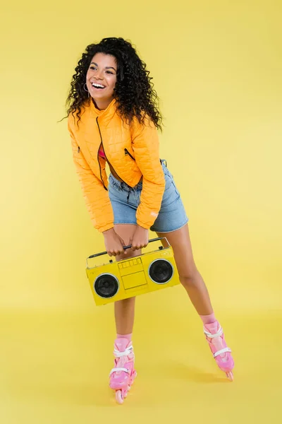 Pleine longueur de femme africaine américaine joyeuse sur patins à roulettes tenant boombox rétro sur jaune — Photo de stock