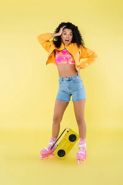 Full length of shocked african american woman on roller skates posing near retro boombox on yellow — Stock Photo