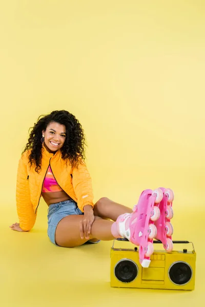 Longitud completa de la alegre mujer afroamericana en patines de ruedas sentado cerca de boombox retro en amarillo - foto de stock