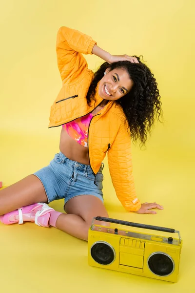 Full length of happy african american woman on roller skates sitting near retro boombox on yellow — Stock Photo