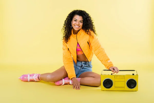 Full length of smiling african american woman on roller skates sitting near retro boombox on yellow — Stock Photo