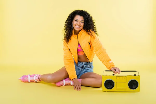 Pleine longueur de souriante femme afro-américaine sur patins à roulettes assis près boombox rétro sur jaune — Photo de stock