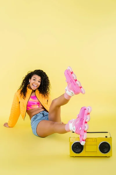 Longitud completa de la mujer afroamericana emocionada en patines de ruedas sentado cerca de boombox retro en amarillo - foto de stock