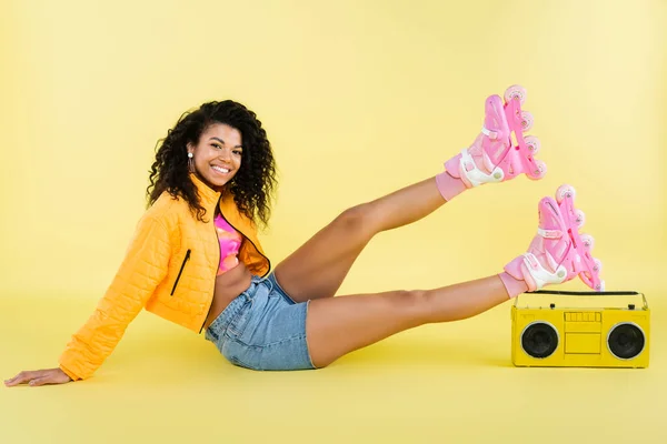 Повна довжина позитивної афроамериканської жінки на роликових ковзанах, що сидять біля ретро бумбоксу на жовтому — стокове фото