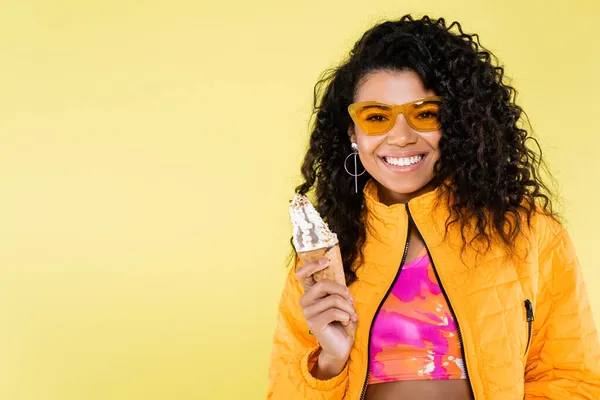 Positivo Africano americano jovem mulher em óculos de sol segurando cone de sorvete isolado em amarelo — Fotografia de Stock