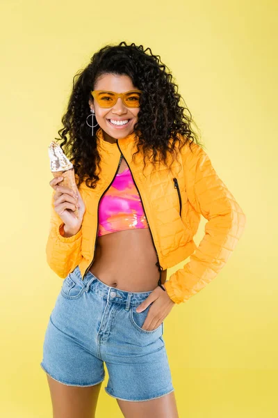 Felice donna afro-americana giovane in occhiali da sole tenendo cono gelato isolato su giallo — Foto stock