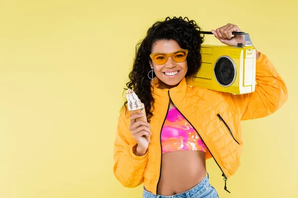 Heureux afro-américaine jeune femme dans des lunettes de soleil tenant cône de crème glacée et boombox isolé sur jaune — Photo de stock