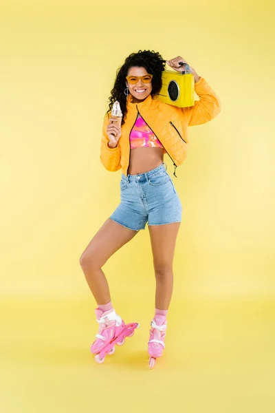 Heureuse jeune femme afro-américaine en patins à roulettes tenant cône de crème glacée et boombox isolé sur jaune — Photo de stock