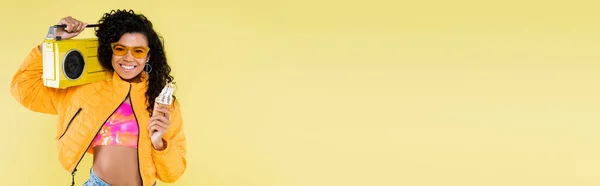Felice donna afroamericana giovane in occhiali da sole che tiene cono gelato e boombox isolato su giallo, banner — Foto stock