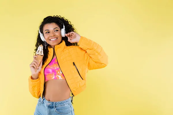 Feliz afro-americana jovem em fones de ouvido segurando cone de sorvete isolado no amarelo — Fotografia de Stock