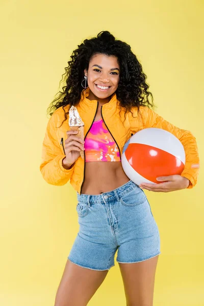 Feliz afro-americana jovem segurando cone de sorvete e bola inflável isolada no amarelo — Fotografia de Stock