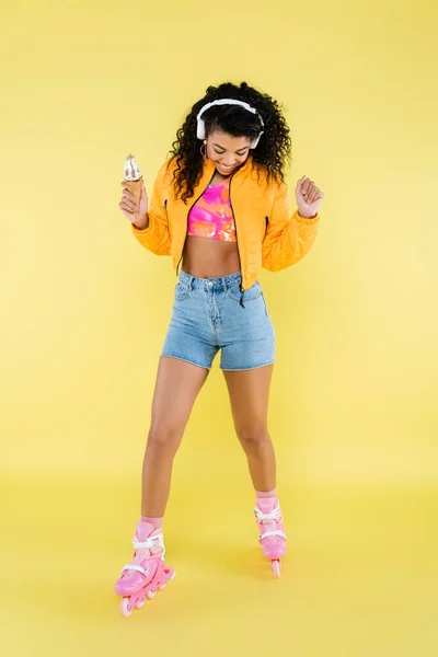 Щаслива афроамериканська молода жінка в навушниках і роликових ковзанах тримає морозиво конус на жовтому — стокове фото