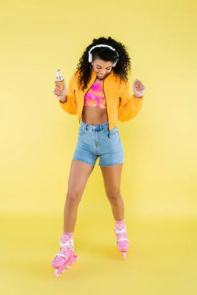 Feliz afro-americana jovem mulher em fones de ouvido e patins segurando cone de sorvete no amarelo — Fotografia de Stock