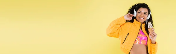 Feliz afro-americana jovem em fones de ouvido segurando cone de sorvete no amarelo, banner — Fotografia de Stock