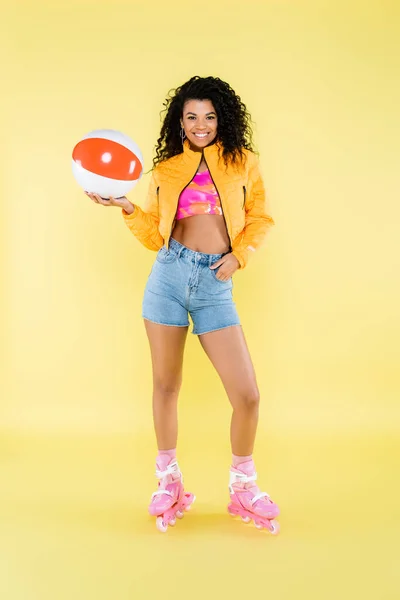 Piena lunghezza della giovane donna afroamericana positiva sui pattini a rotelle che tiene palla gonfiabile sul giallo — Foto stock