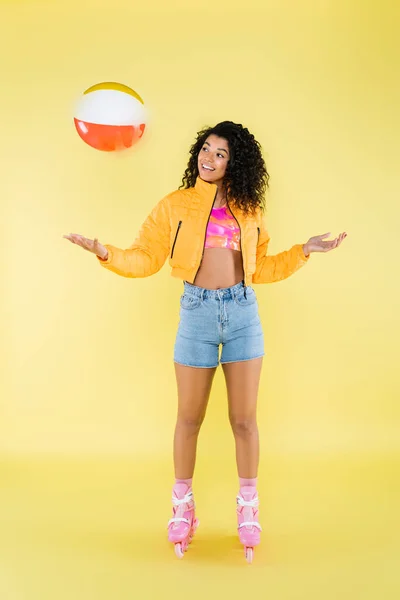 Pleine longueur de jeune femme afro-américaine positive en patins à roulettes jetant ballon gonflable sur jaune — Photo de stock