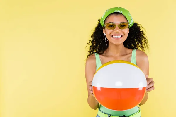 Mujer joven afroamericana positiva en pañuelo sosteniendo bola inflable aislada en amarillo - foto de stock