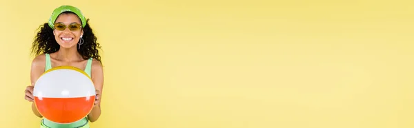 Positivo afroamericano giovane donna in fazzoletto e occhiali da sole tenendo palla gonfiabile isolato su giallo, banner — Foto stock