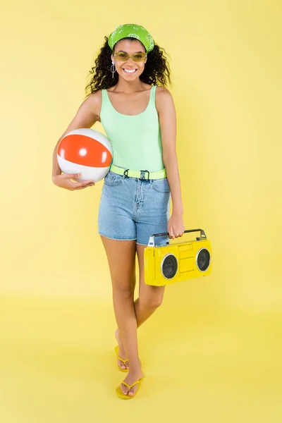 Volle Länge der glücklichen afrikanisch-amerikanischen Frau in Sonnenbrille mit Boombox und aufblasbarem Ball auf gelb — Stockfoto