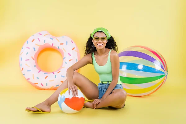 Pleine longueur de femme afro-américaine heureuse assise avec balle de plage sur jaune — Photo de stock