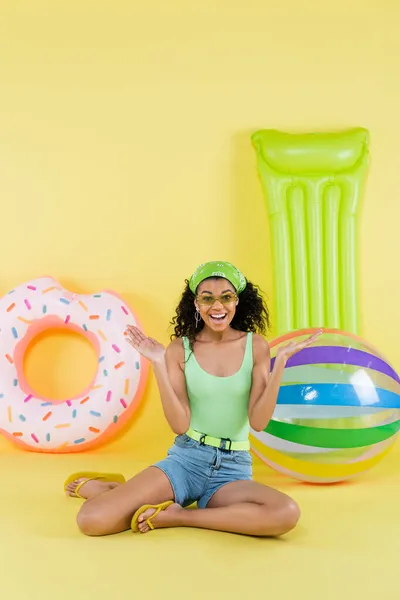 Full length of cheerful african american woman sitting near inflatable ball, mattress and ring on yellow — Stock Photo