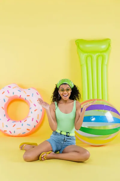 Pleine longueur de femme américaine africaine joyeuse assise près de ballon gonflable, matelas et anneau sur jaune — Photo de stock