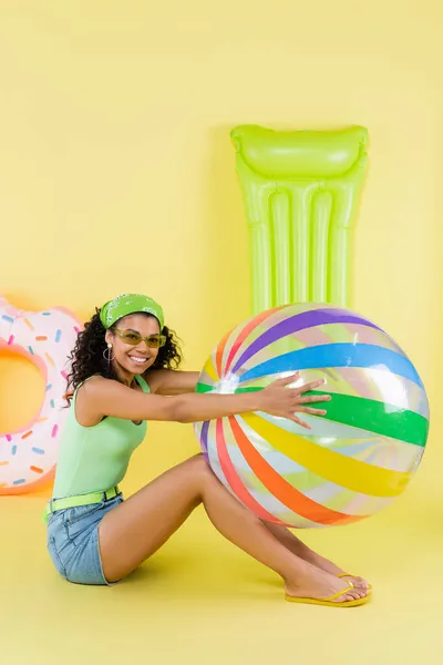 Full length of joyful african american woman sitting with inflatable ball, mattress and ring on yellow — Stock Photo