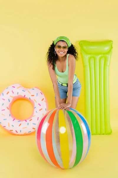 Pleine longueur de femme africaine américaine positive près de ballon gonflable, matelas et anneau sur jaune — Photo de stock