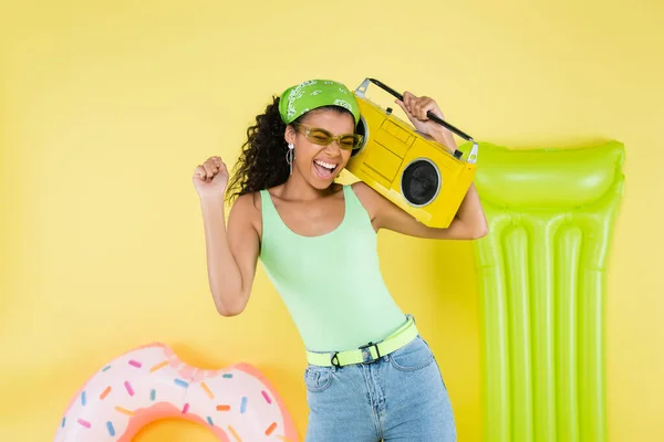 Cheerful african american young woman holding boombox near inflatable mattress and ring on yellow — Stock Photo
