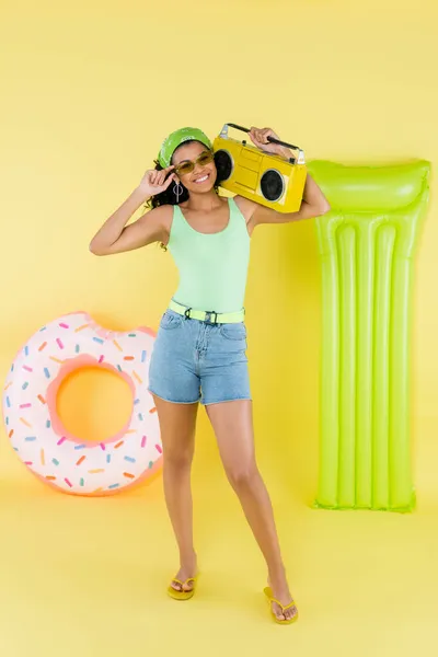 Full length of cheerful african american young woman holding boombox near inflatable mattress and ring on yellow — Stock Photo