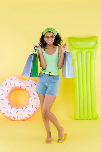 Full length of cheerful african american young woman holding shopping bags near inflatable mattress and ring on yellow — Stock Photo
