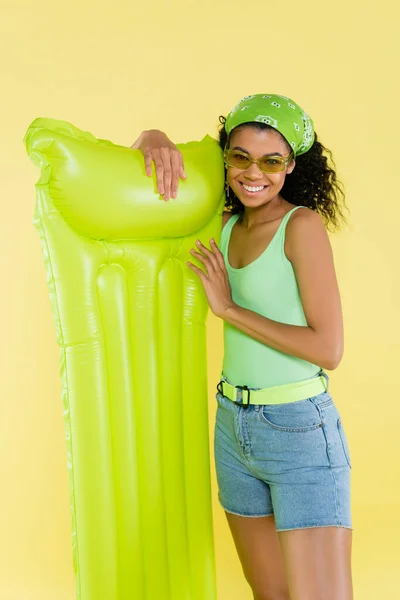 Весела афроамериканська молода жінка, що стоїть з надувним матрацом ізольована на жовтому — стокове фото