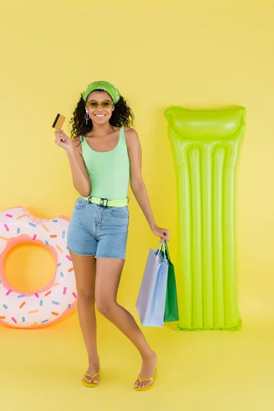 Full length of cheerful african american woman holding shopping bags and credit card near inflatable mattress and ring on yellow — Stock Photo