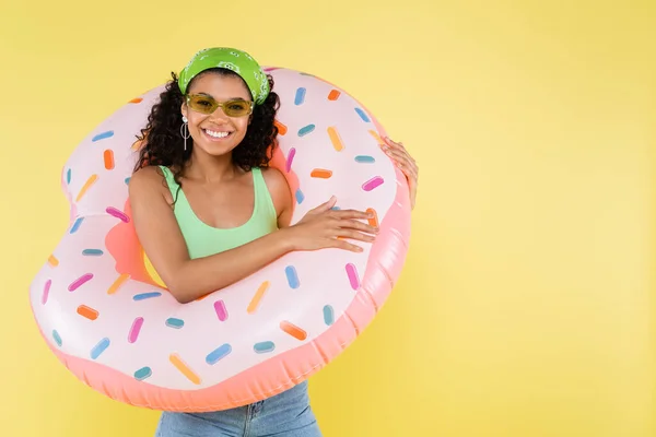 Positive afro-américaine jeune femme debout avec anneau gonflable isolé sur jaune — Photo de stock