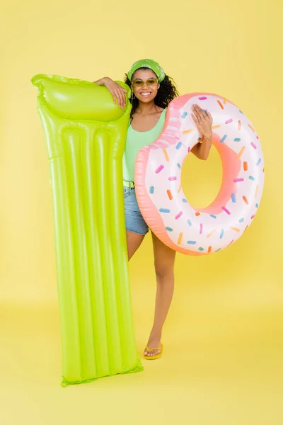 Full length of happy african american woman holding inflatable mattress and ring on yellow — Stock Photo