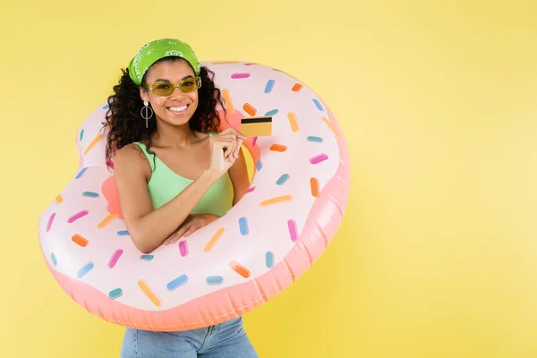 Mujer joven afroamericana positiva de pie con anillo inflable y tarjeta de crédito aislada en amarillo - foto de stock