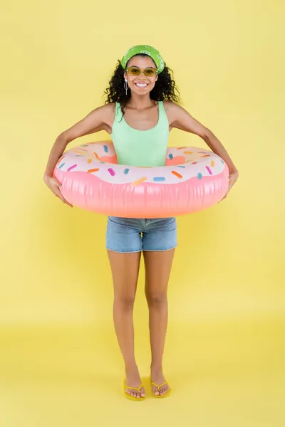 Piena lunghezza della giovane donna afroamericana gioiosa in piedi con anello gonfiabile sul giallo — Foto stock