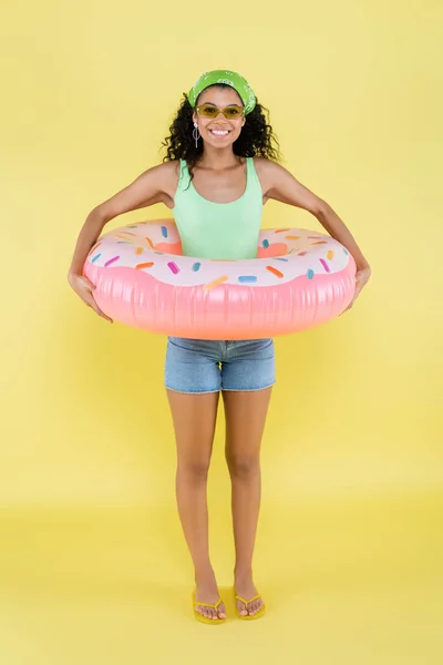 Longitud completa de la joven afroamericana alegre que está parada con el anillo inflable en amarillo - foto de stock