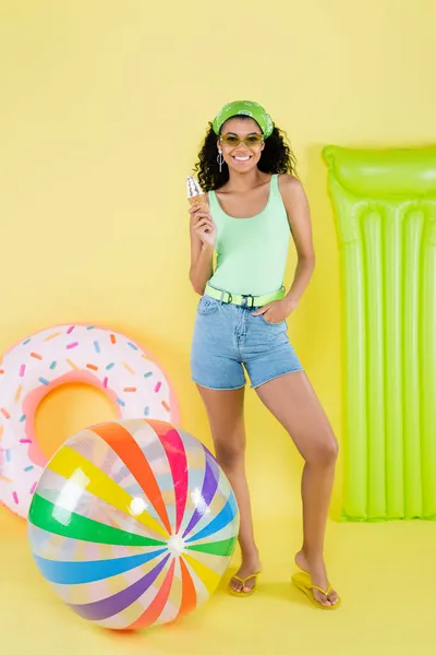 Pleine longueur de jeune femme africaine américaine positive debout avec cône de crème glacée près ballon gonflable, matelas et anneau sur jaune — Photo de stock