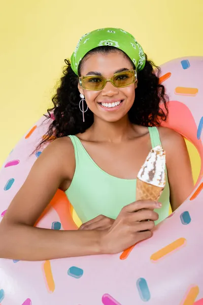 Gioiosa giovane donna afroamericana in piedi con anello gonfiabile e cono gelato isolato su giallo — Foto stock