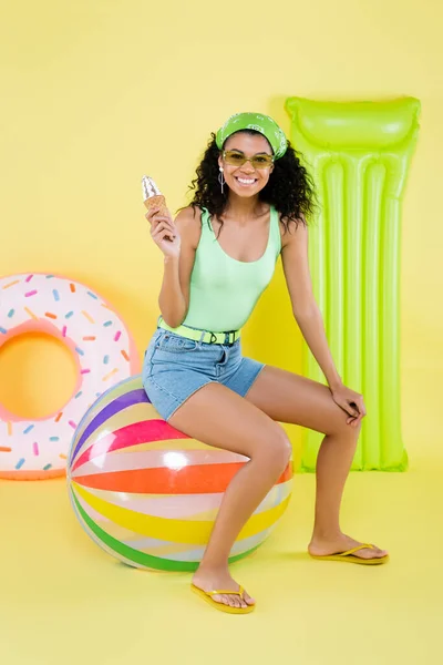 Full length of positive african american young woman sitting on beach ball and holding ice cream near inflatable mattress and ring on yellow — Stock Photo