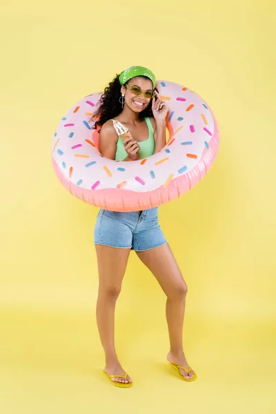 Piena lunghezza di gioiosa giovane donna africana americana in piedi con anello gonfiabile e cono gelato su giallo — Foto stock
