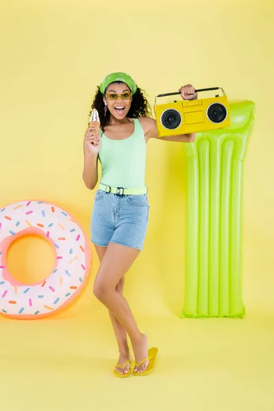 Pleine longueur de joyeuse jeune femme afro-américaine debout avec boombox et cône de crème glacée près anneau gonflable et matelas sur jaune — Photo de stock