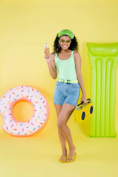 Pleine longueur de jeune femme afro-américaine heureuse debout avec boombox et cône de crème glacée près anneau gonflable et matelas sur jaune — Photo de stock