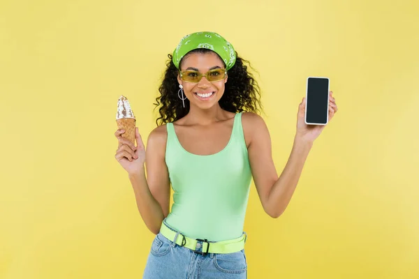 Happy african american woman in sunglasses holding ice cream cone and smartphone with blank screen isolated on yellow — Stock Photo