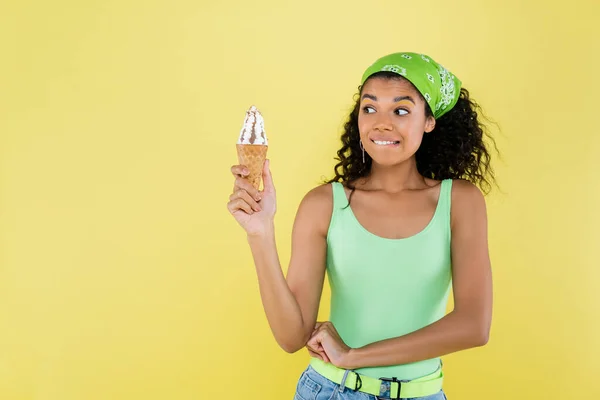 Encaracolado afro-americano mulher em lenço verde segurando sorvete e mordendo lábios isolados no amarelo — Fotografia de Stock