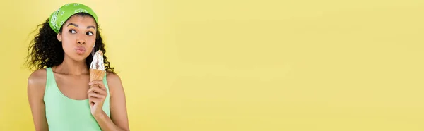 Femme afro-américaine bouclée en mouchoir vert tenant crème glacée et lèvres boudantes isolées sur jaune, bannière — Photo de stock
