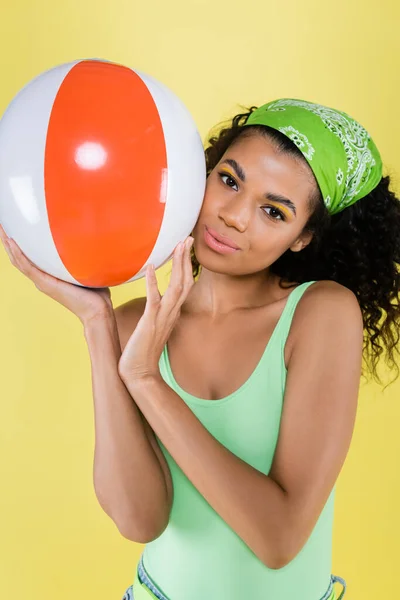 Joven afroamericana joven mujer en verde pañuelo celebración inflable bola aislado en amarillo - foto de stock